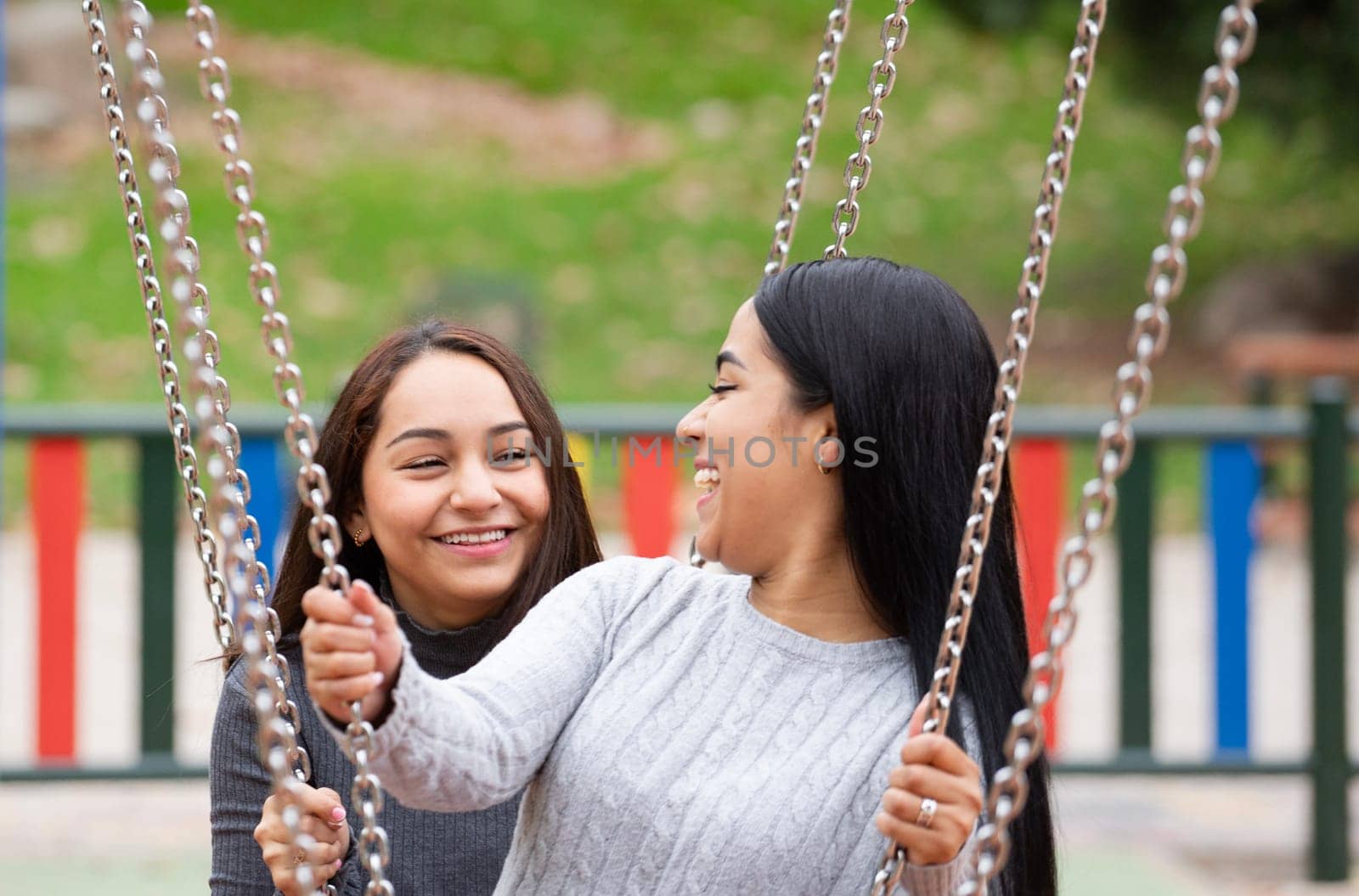 Portrait of two latin women friends are swinging on a swing set, smiling and laughing.