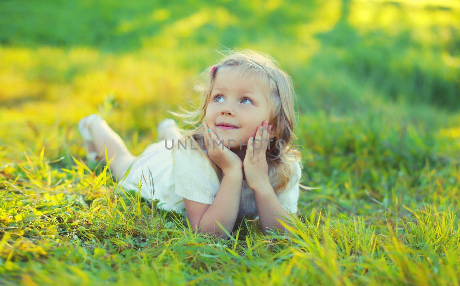 Happy little girl child lying on green grass looking up in sunny summer park by Rohappy