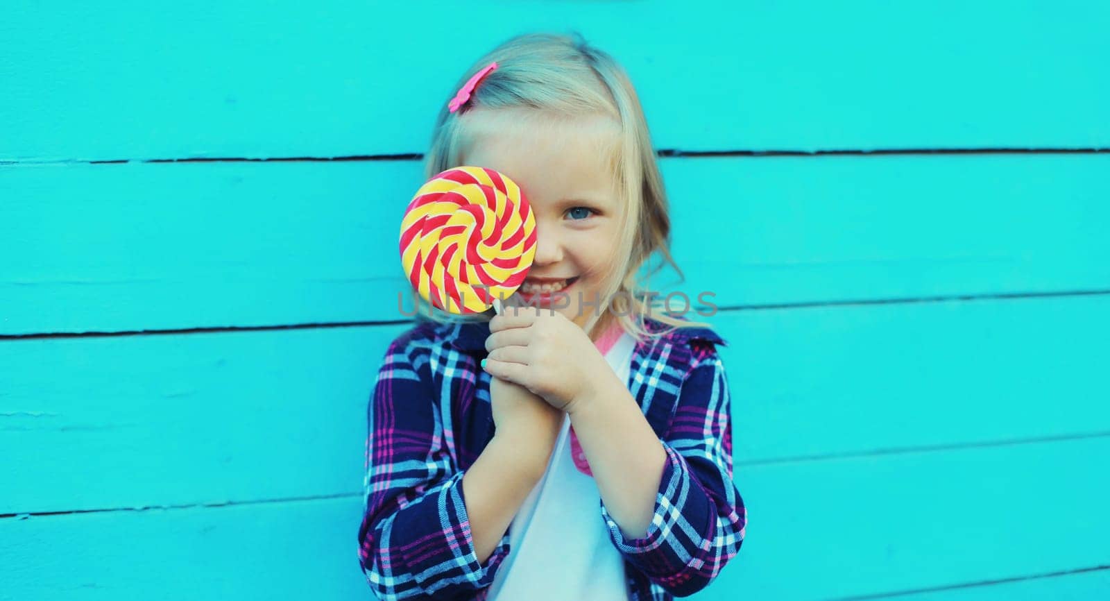 Portrait of happy cheerful smiling little girl child with sweet lollipop on stick on blue background