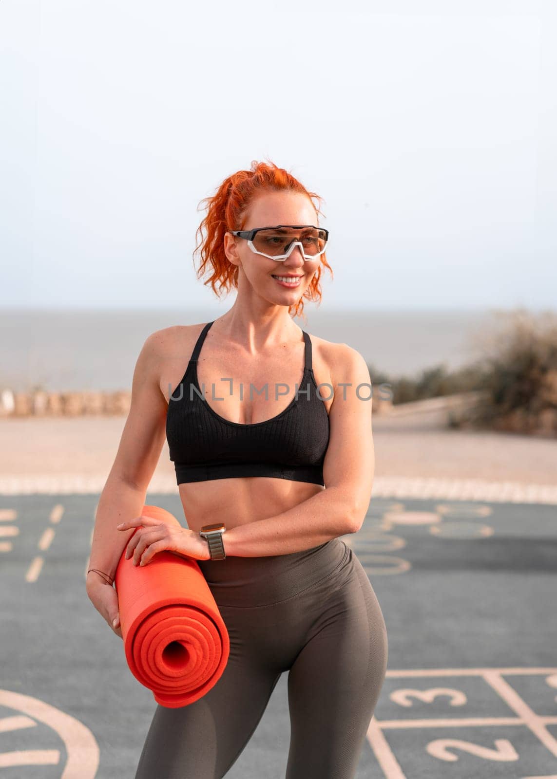 Smiling redhead woman in sunglasses holding rolled yoga mat and looking away. Fit sporty female athlete in activewear looking away at summer park. Fitness and health concept.