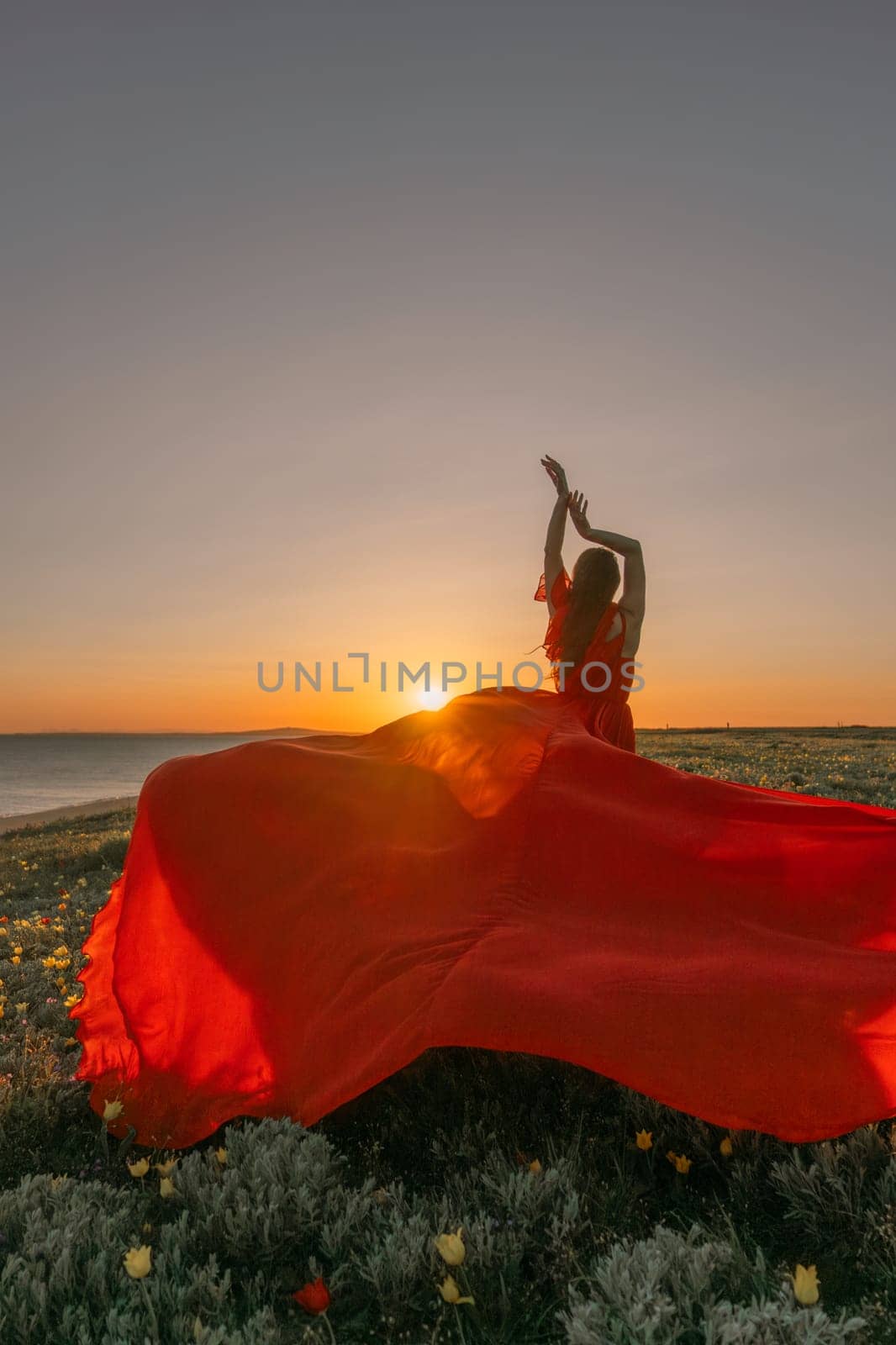 A woman in a red dress is standing in a field with the sun setting behind her. She is reaching up with her arms outstretched, as if she is trying to catch the sun. The scene is serene and peaceful. by Matiunina