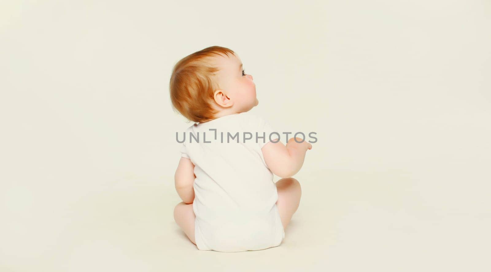 Happy cute baby sitting on the floor on white studio background, looking up, back view by Rohappy