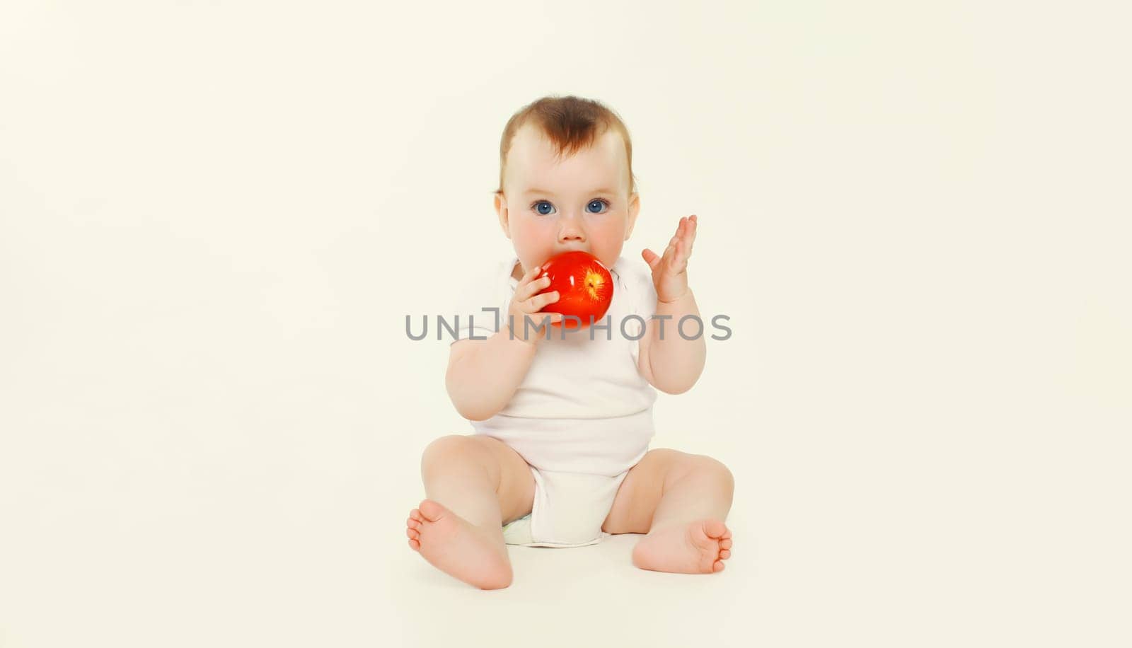 cute baby holding red apple fruit sitting on the floor on white studio background by Rohappy