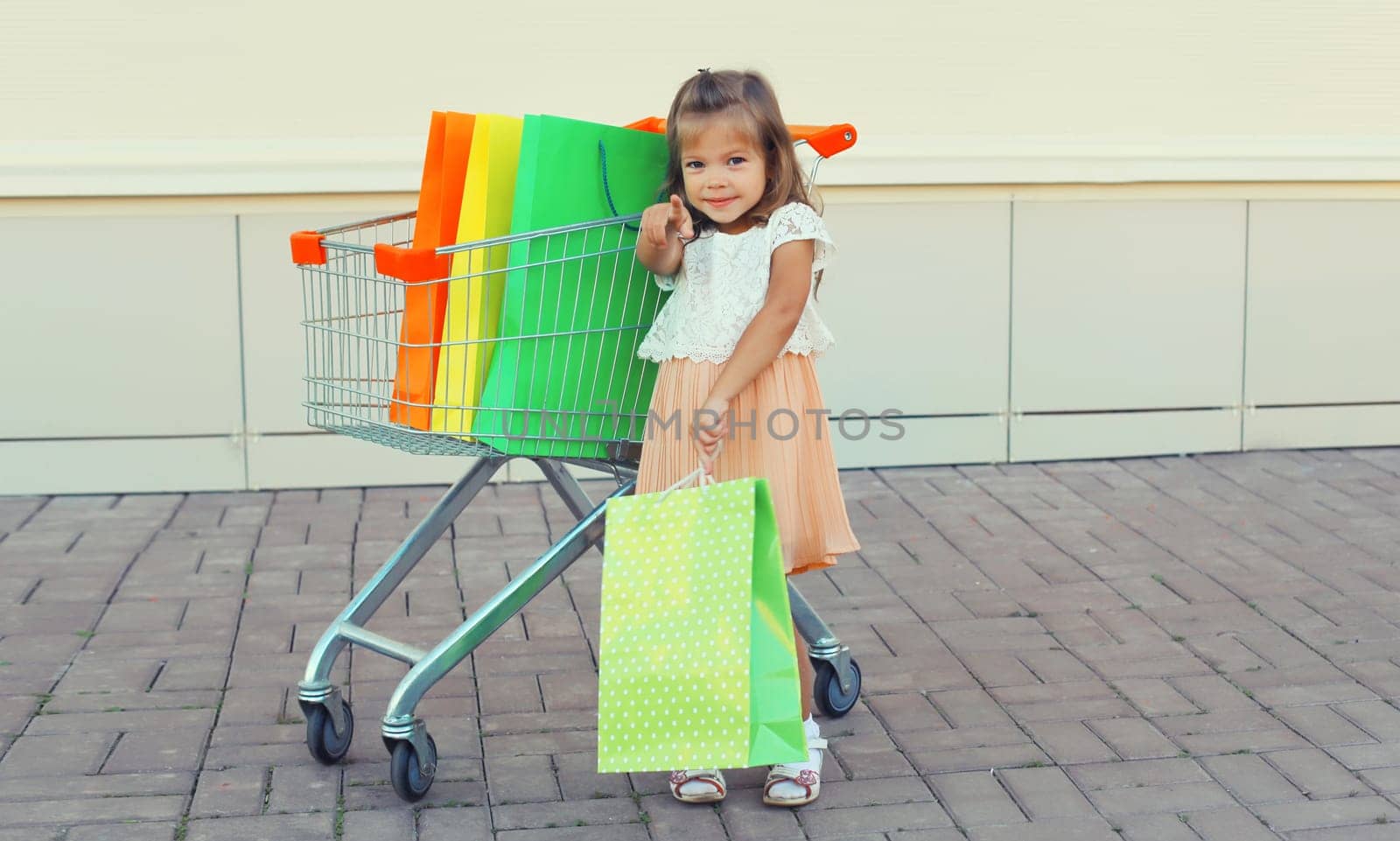 Happy little girl child with trolley cart and colorful shopping bags posing on city street