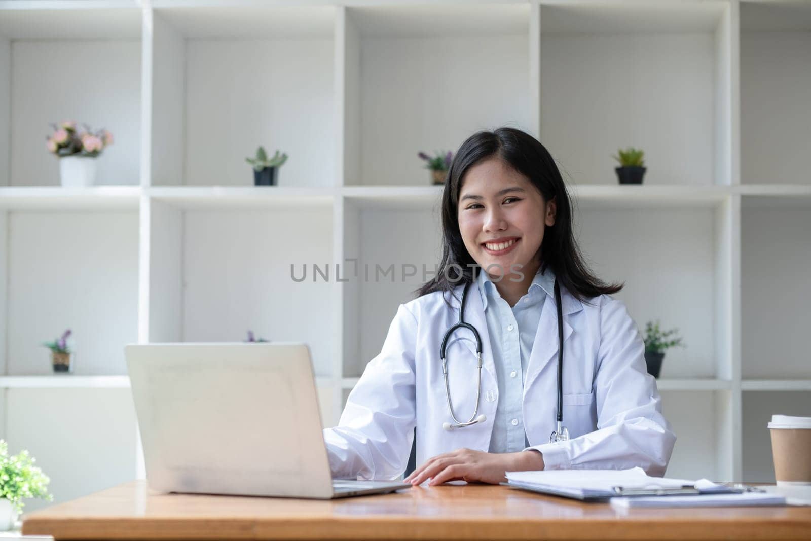 young asian lady doctor in white medical uniform with stethoscope using computer laptop talking video conference call with patient at desk in health clinic or hospital. by wichayada