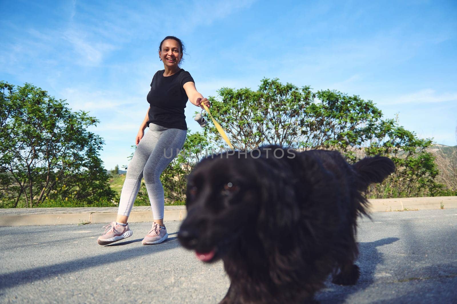 Happy woman walking her dog outdoors. Pretty female athlete dressed in gray leggings and black t-shirt, walking her cocker spaniel pet on leash by artgf