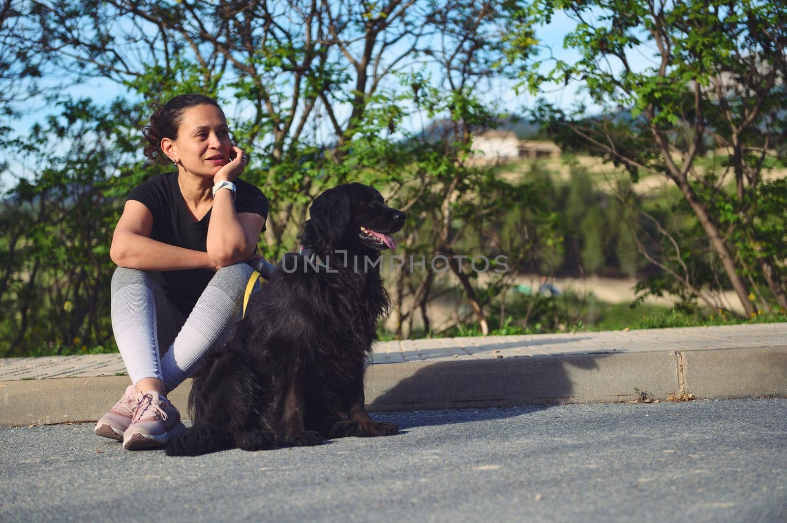 Multi ethnic beautiful pensive woman sitting next to her dog on the asphalt, a beautiful black cocker spaniel and looking dreamily into the distance. Pets and people. Woman walking her dog on leash by artgf