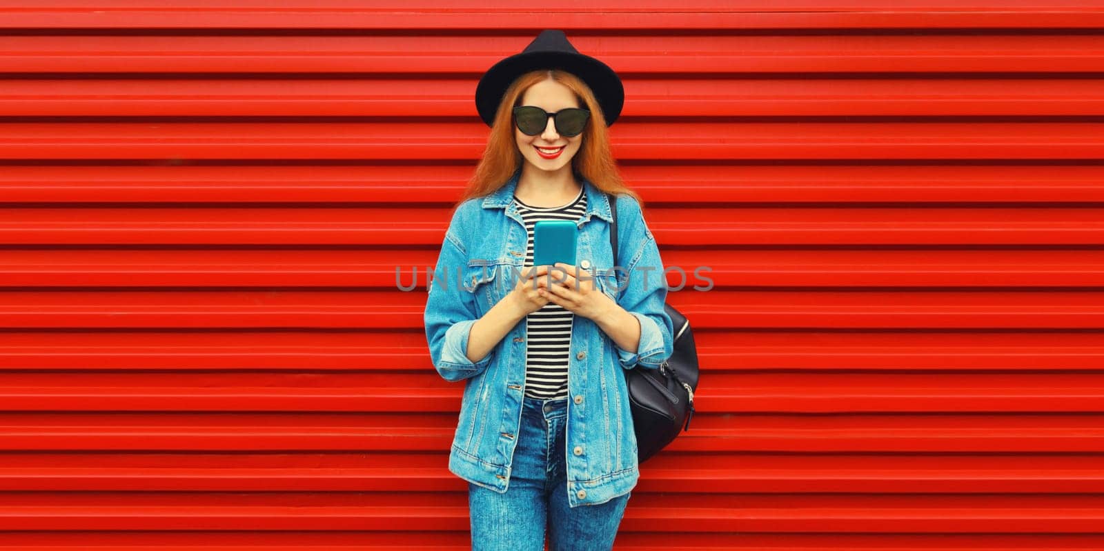 Portrait of stylish modern smiling young woman with smartphone wearing jean jacket, black round hat on red background
