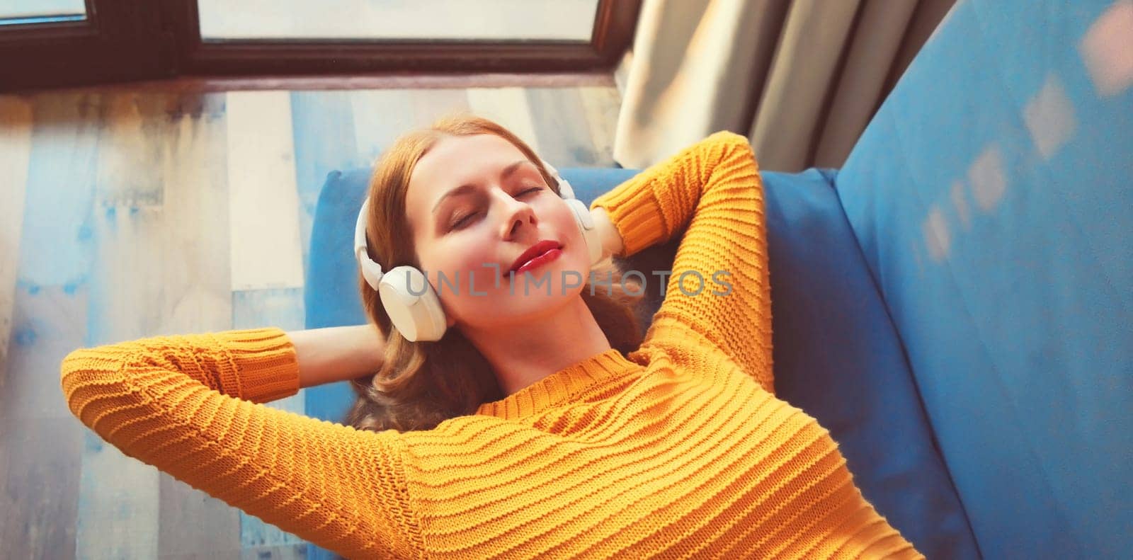 Happy relaxed young woman listening to music with wireless headphones with closed eyes lies on the couch at home
