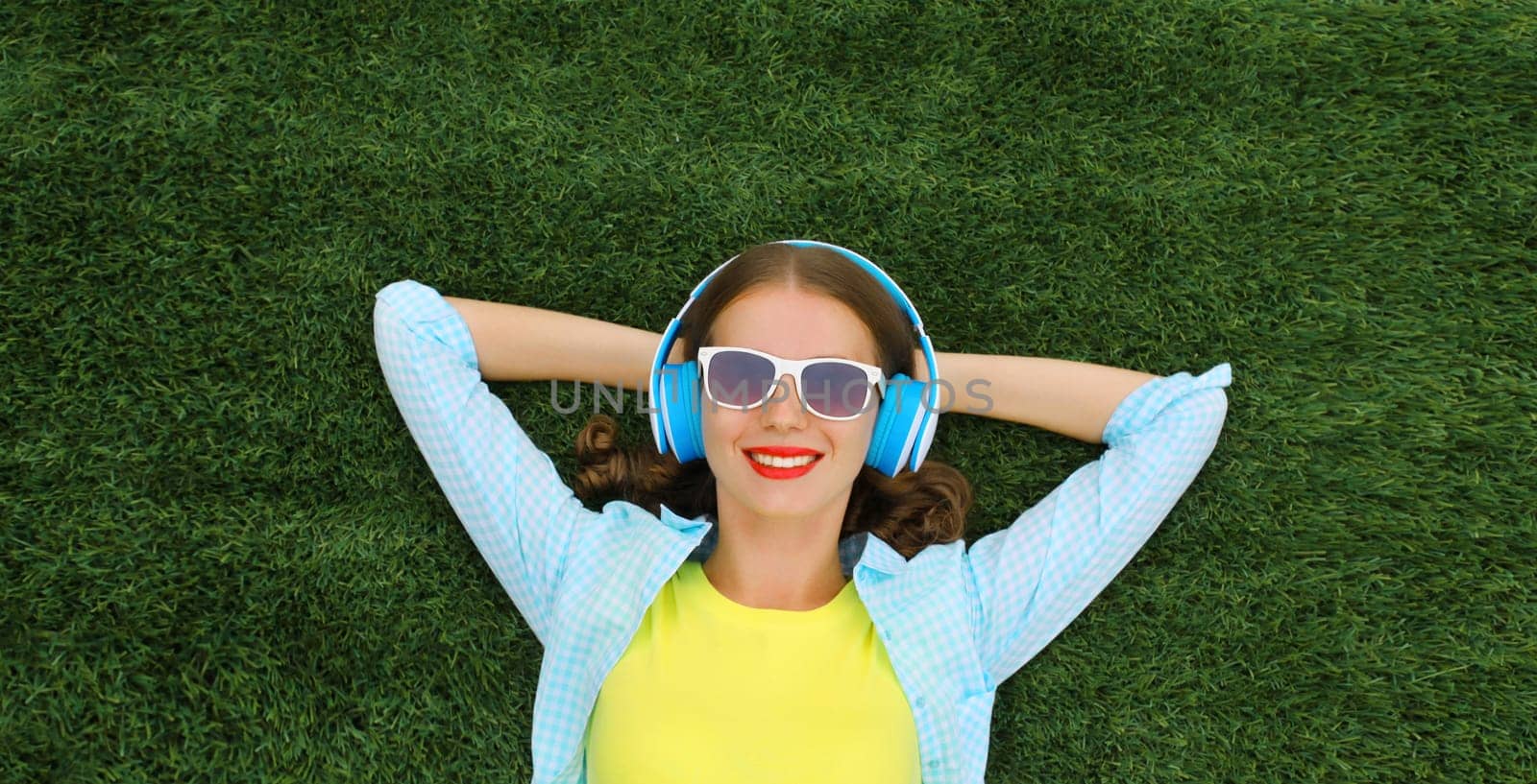 Happy smiling young woman listening to music in headphones while lying on grass in summer park by Rohappy