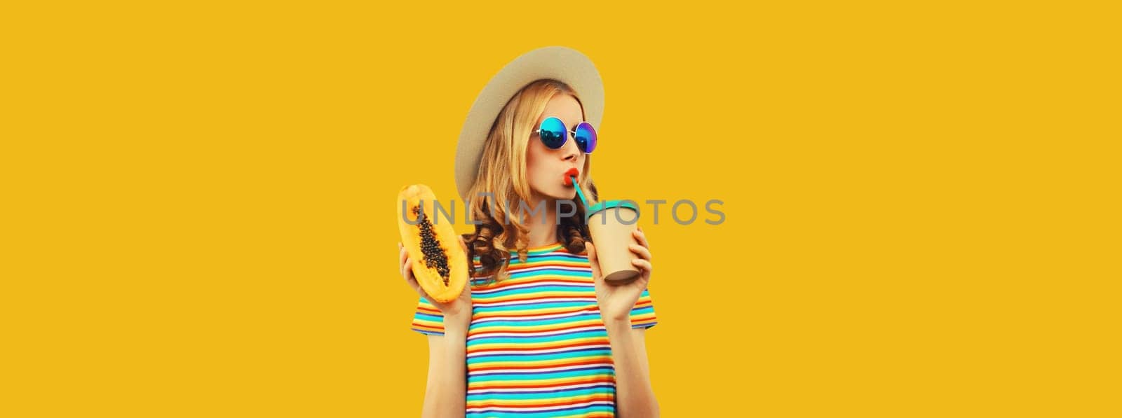 Summer portrait of stylish young woman with juicy papaya fruit drinking fresh juice looking away in straw hat, sunglasses on colorful yellow studio background