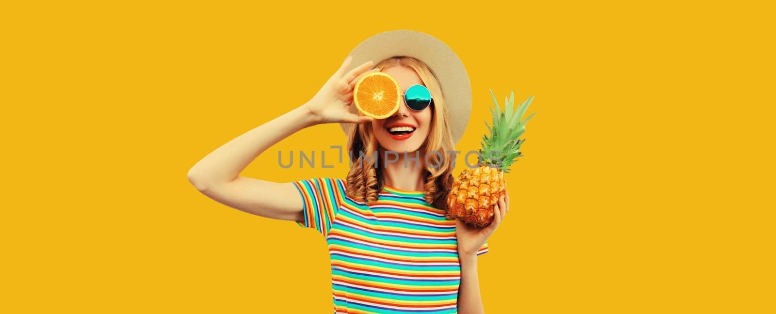 Summer vacation portrait of happy smiling young woman with fresh tropical juicy fruits, pineapple, slice of orange in straw tourist hat on yellow studio background