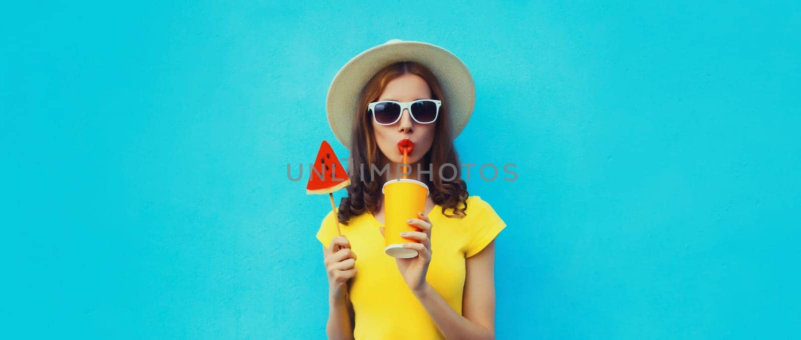 Summer portrait of young woman drinking juice with sweet juicy lollipop or ice cream shaped slice of watermelon wearing straw hat on blue background