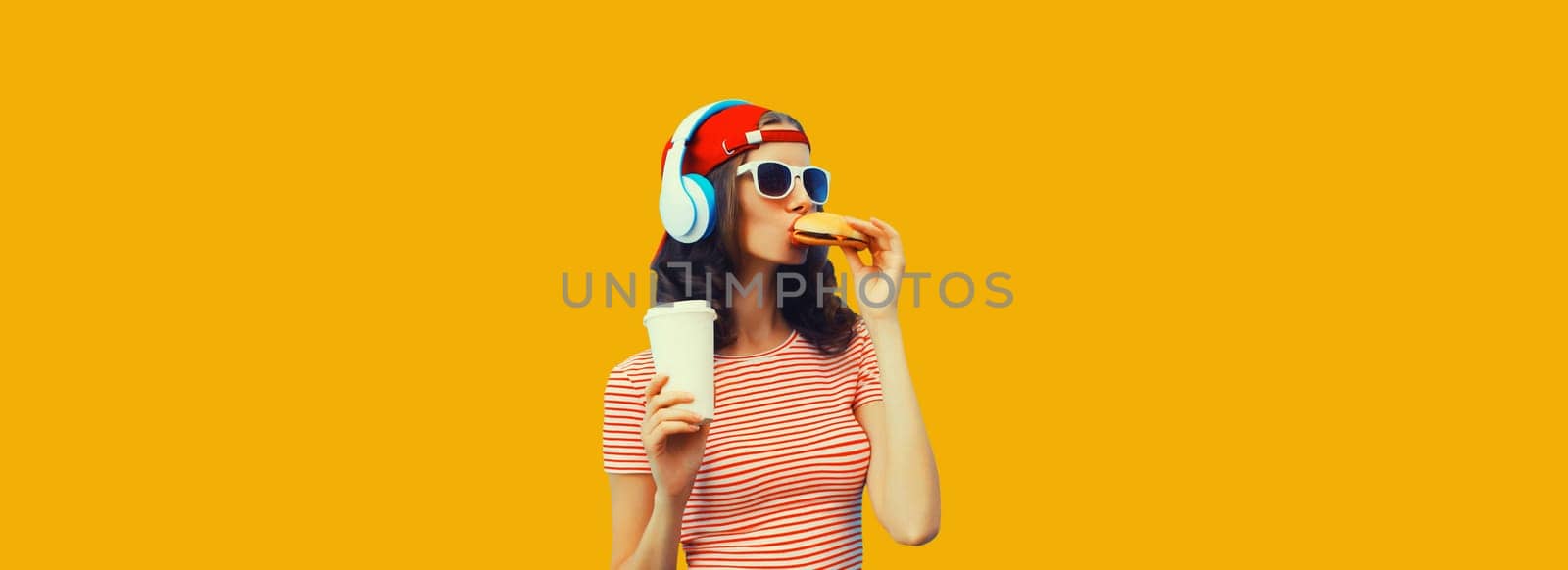 Stylish young woman listening to music in headphones eating burger fast food holding cup of coffee or juice on yellow background