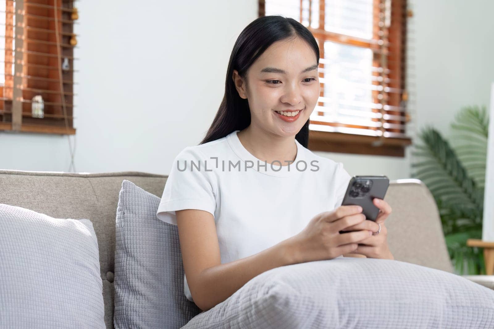 Happy young asian woman relax on comfortable couch at home texting messaging on smartphone, shopping online from home.