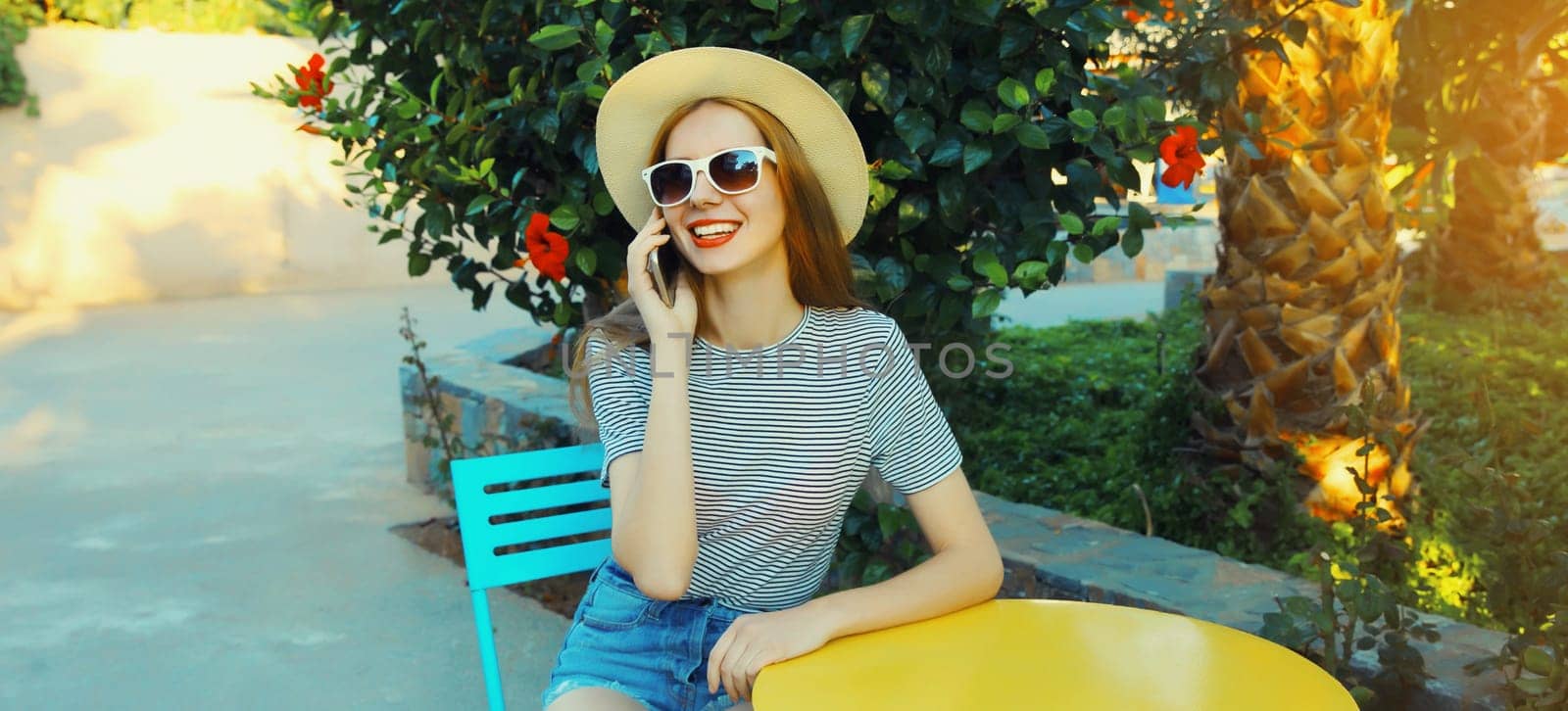 Happy modern smiling young woman calling on mobile phone sitting on the table waiting in street cafe in summer park