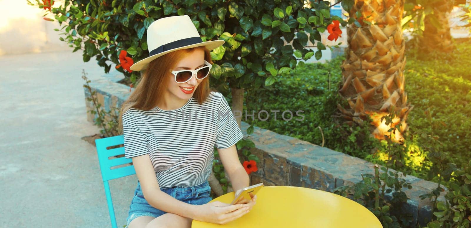 Happy caucasian young woman 20s with mobile phone looking at device sitting at the table in street cafe in summer park