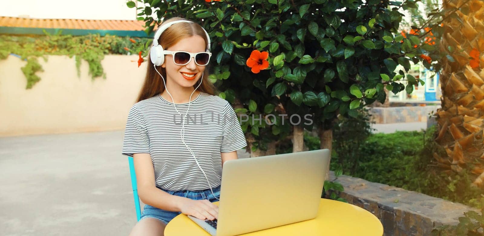 Happy young woman working with laptop listening to music in headphones sitting at the table in street cafe in summer park