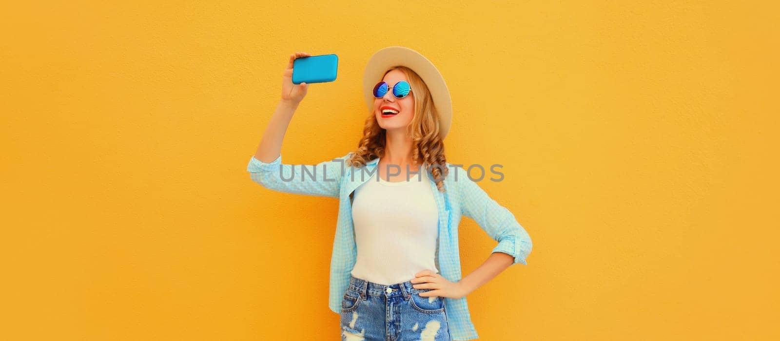 Portrait of happy smiling young woman taking selfie with smartphone wearing straw hat on yellow background