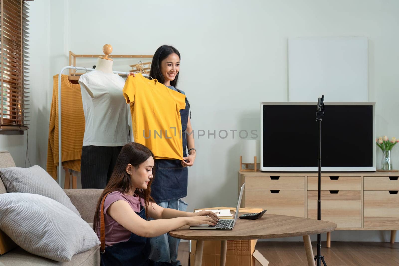 Asian woman friends couple talk to camera to sell the clothes at home. Young attractive business people female live streaming and showing clothing to customer in online.