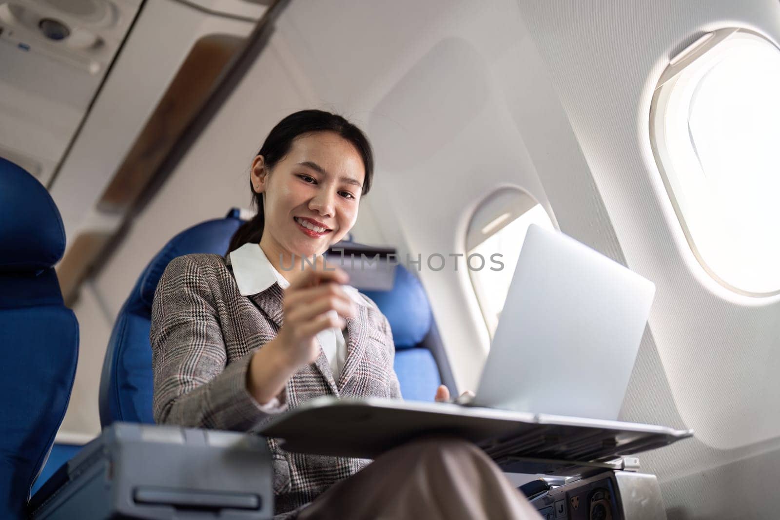 Young Asian businesswoman holding credit card in online shopping on laptop sitting near window on airplane during flight, travel and business tourism concept by nateemee