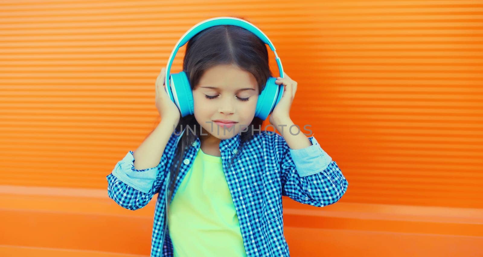 Portrait of happy little girl child in blue wireless headphones listening to music on orange background