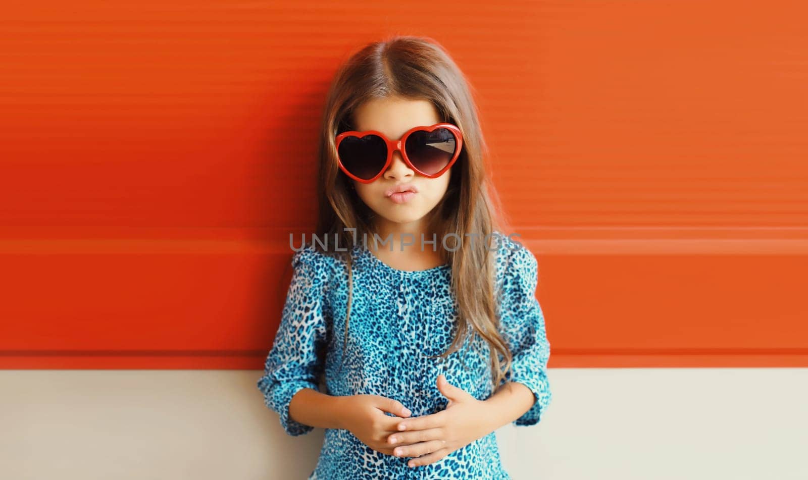 Portrait of beautiful little girl child in red heart shaped sunglasses and blue dress on city street