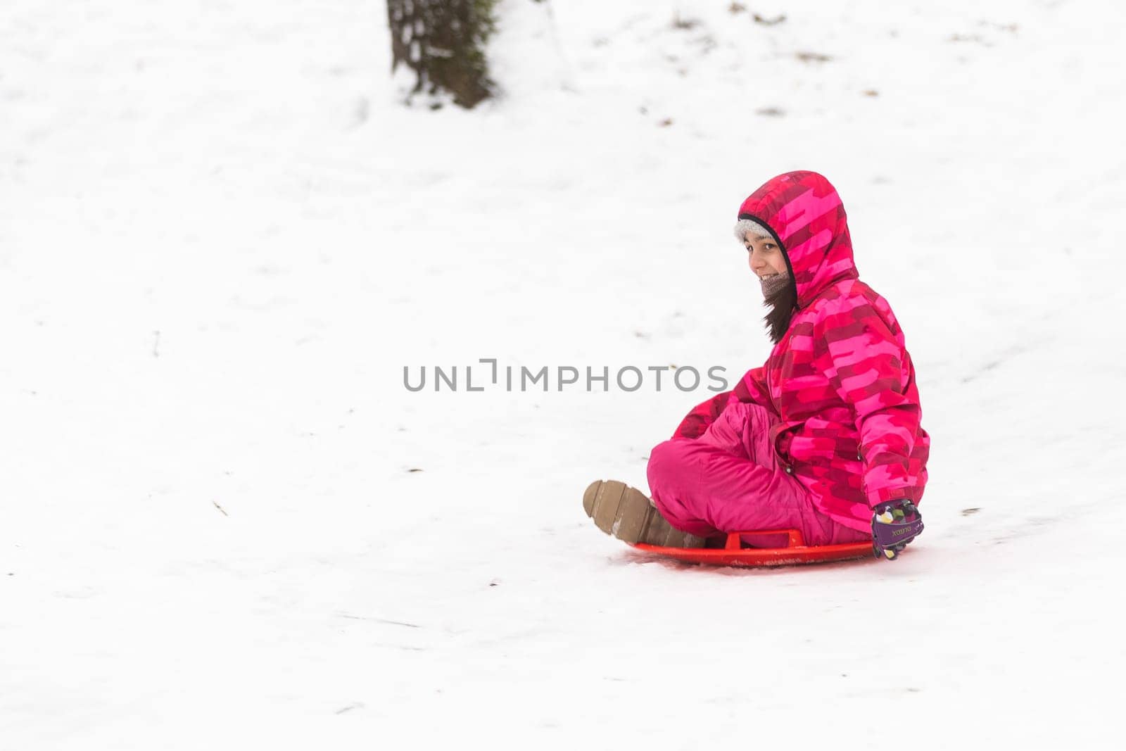 Pretty little girl wearing pink jacket and knitted hat walking in snowy winter park. Child playing with ice skating plate in forest. Family vacation with child in mountains by Andelov13