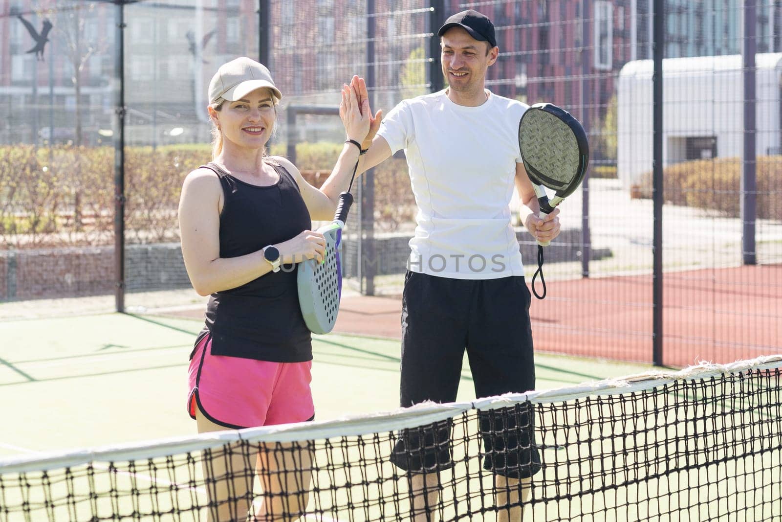 Portrait of positive young woman and adult man standing on padel tennis court, holding racket and ball, smiling by Andelov13