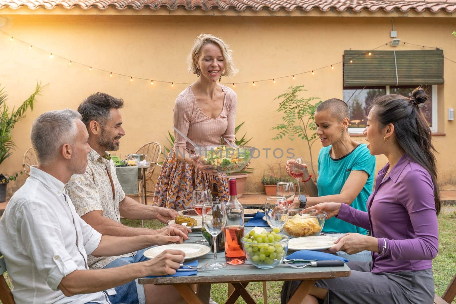Multiethnic friends laughing with big smile around the table. Woman serves the food at house patio. Smiles, cheerfulness concept. by PaulCarr