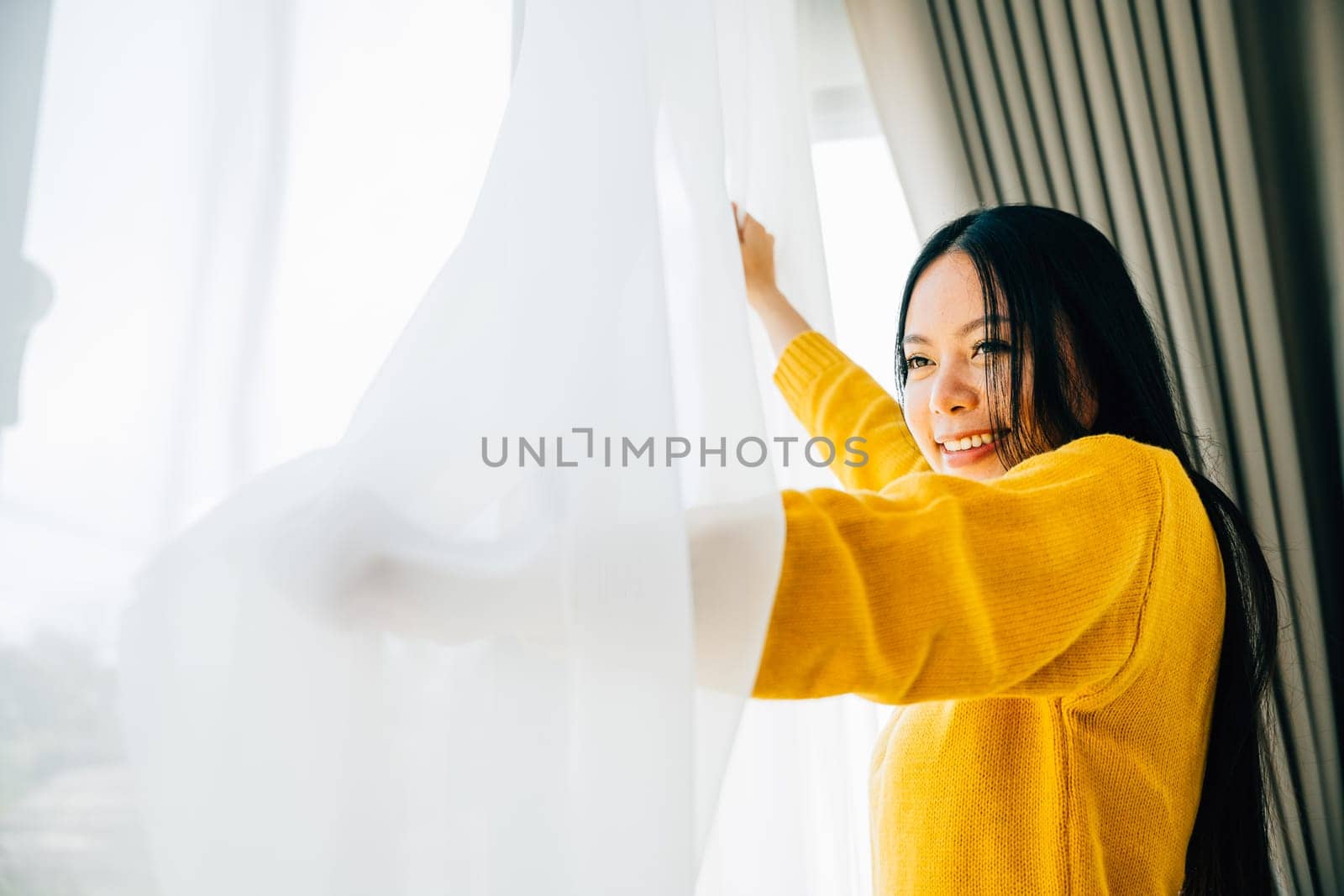 A young woman opens curtains in the early morning smiles at the view feeling relaxed by Sorapop