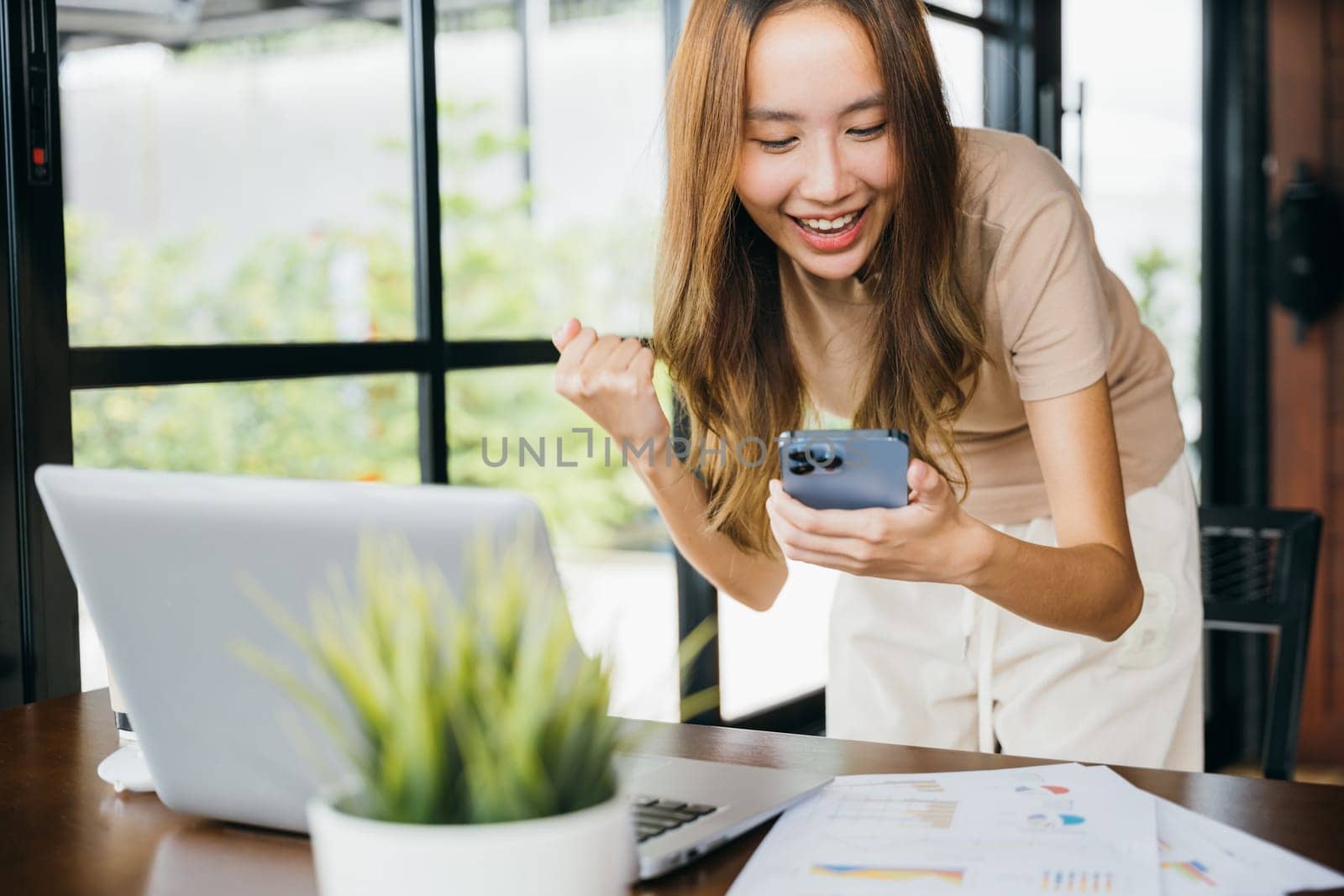 Excited Asian young woman holding mobile smart phone celebrate with laptop, happy success, female got good news on smartphone while working on computer at cefa, social media network