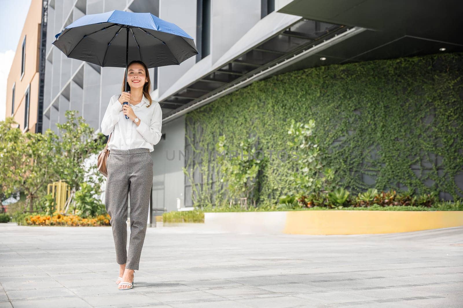 Walking to the office on a hot day, a young businesswoman holds an umbrella to protect herself from the sun rays. Her professional demeanor and makeup reflect her determination and success. by Sorapop