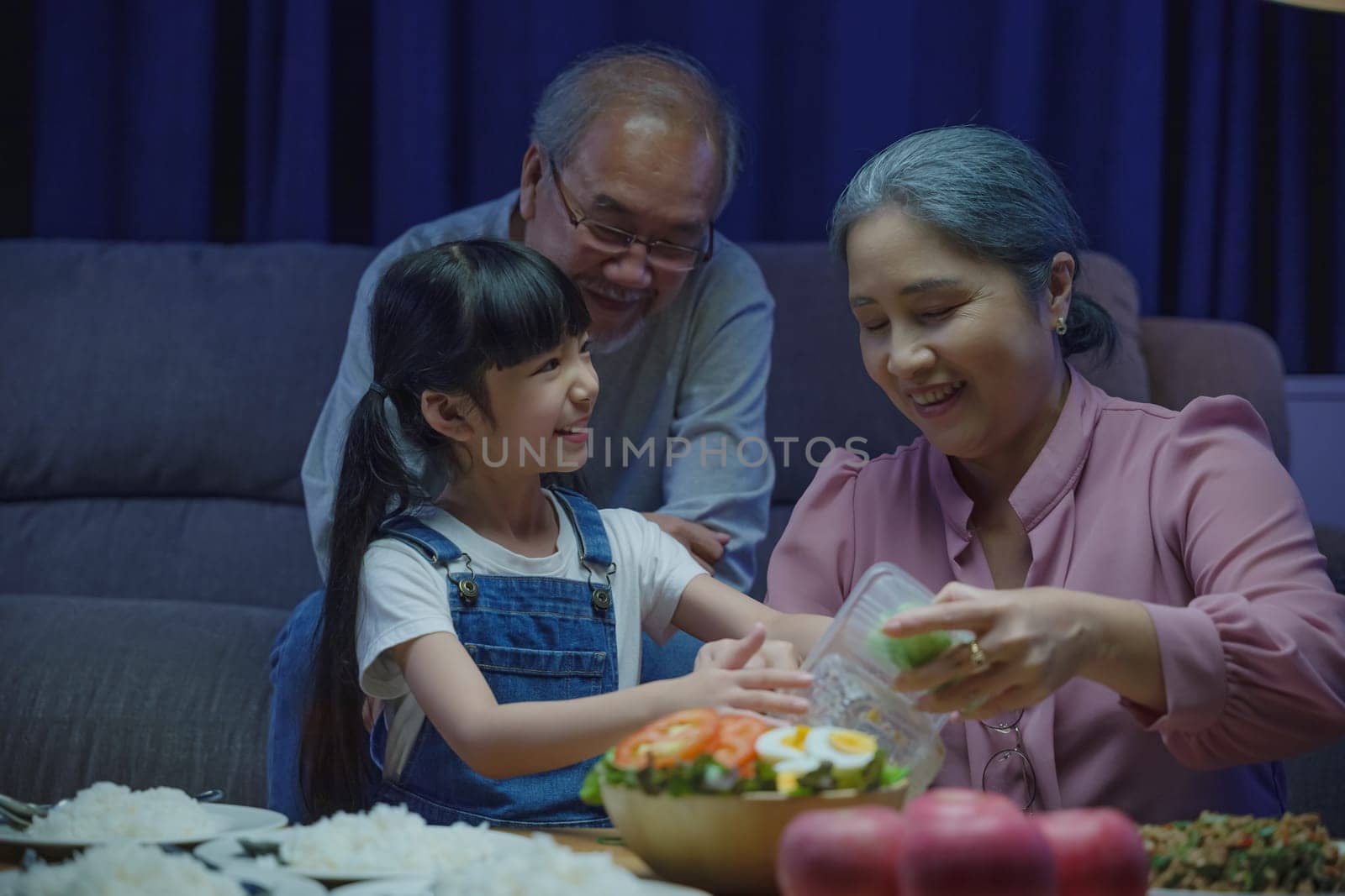 Happy Asian senior parent and child eating dinner food together in living room indoors, family grandmother grandfather and granddaughter dining on table and having fun during at home night time