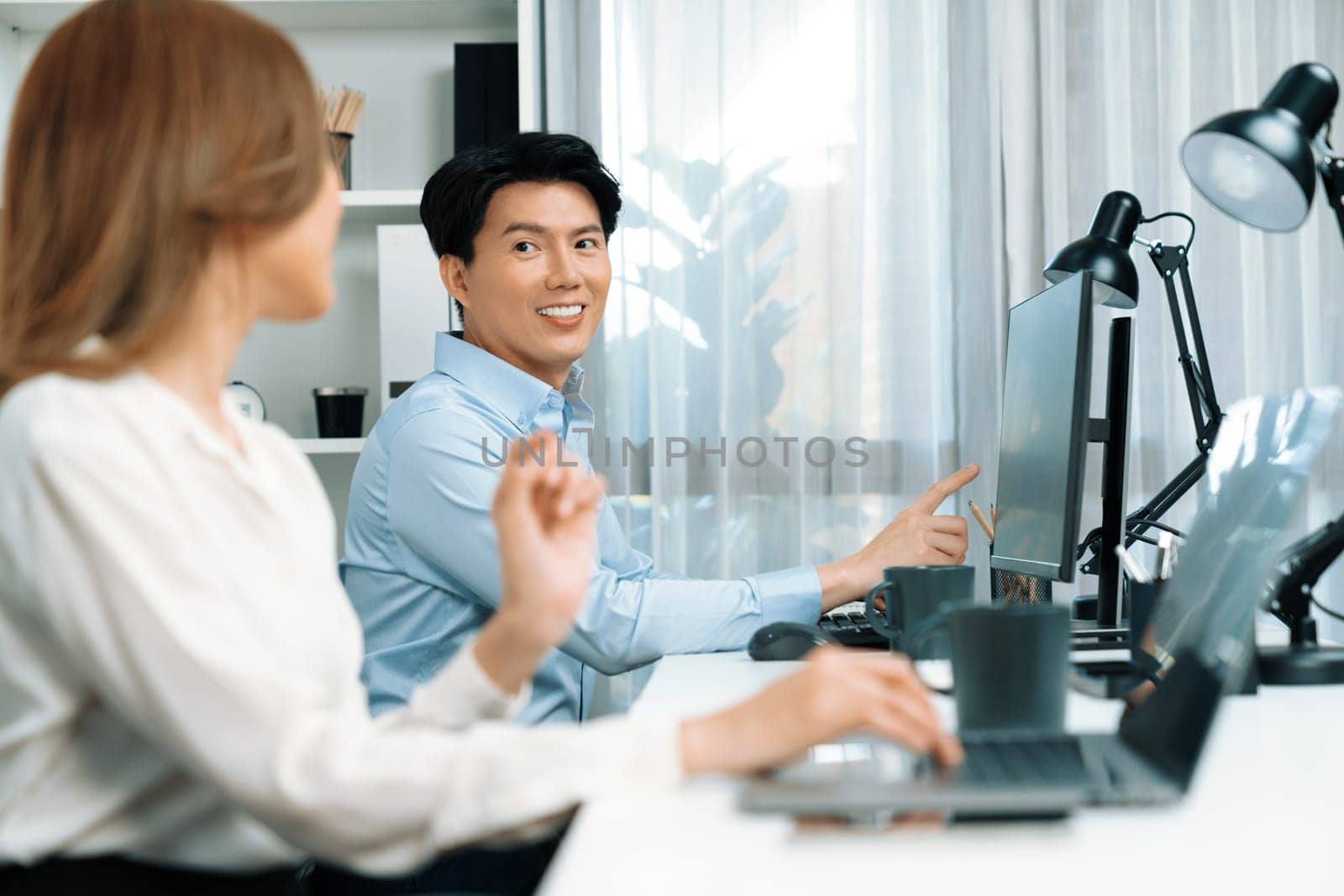 Smiling Asian man talking with woman colleague in planning marketing report project on laptop casual day while searching database on working desk at modern home office at side view image. Infobahn.