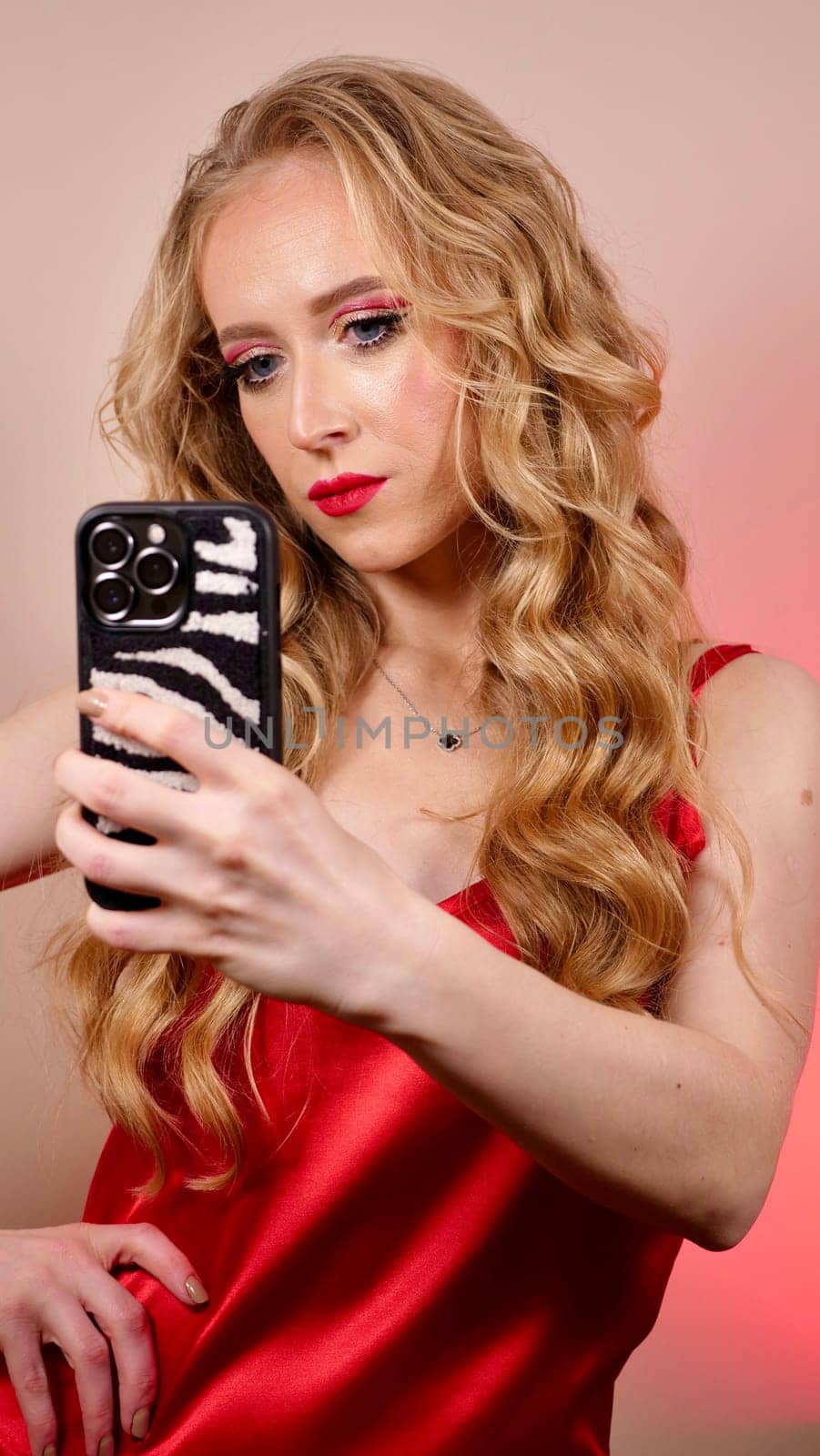 A young woman with red lipstick takes a selfie with long blond hair and makeup. The girl is smiling and looking at the camera close-up