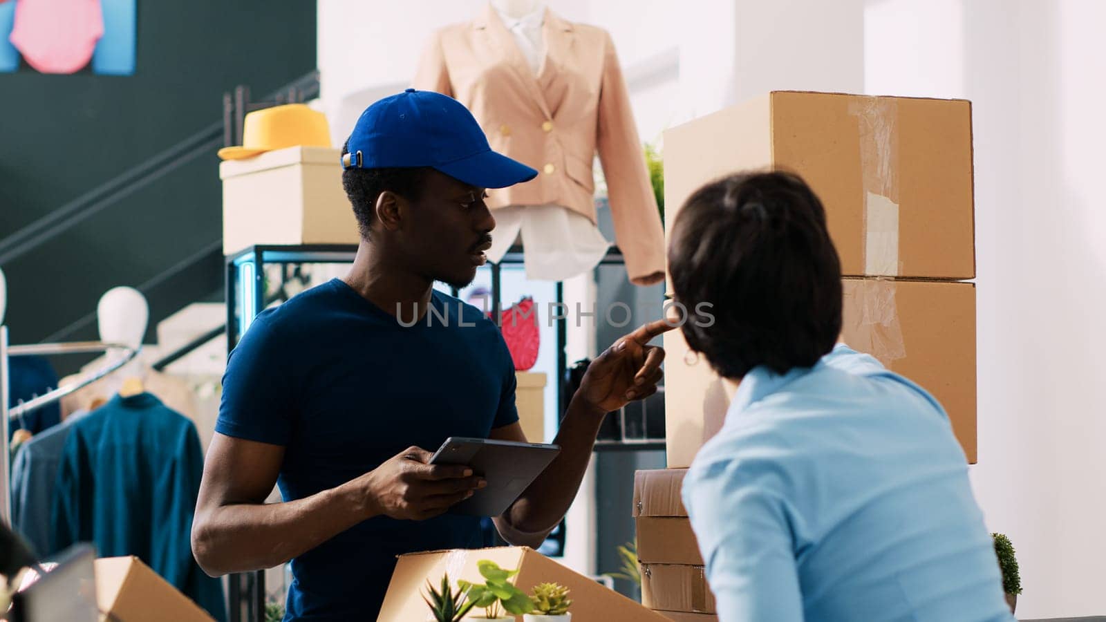 Store employee signing shipping report, discussing logistics with african american courier in shopping centre. Stylish manager preparing customers packages for delivery in modern boutique