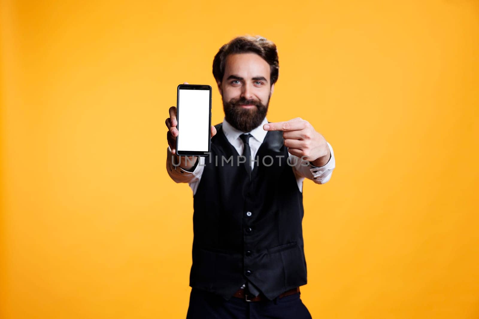 Confident waiter points to white screen on camera, presenting isolated mockup blank display on mobile phone. Young butler with suit and tie shows empty device with copyspace template.