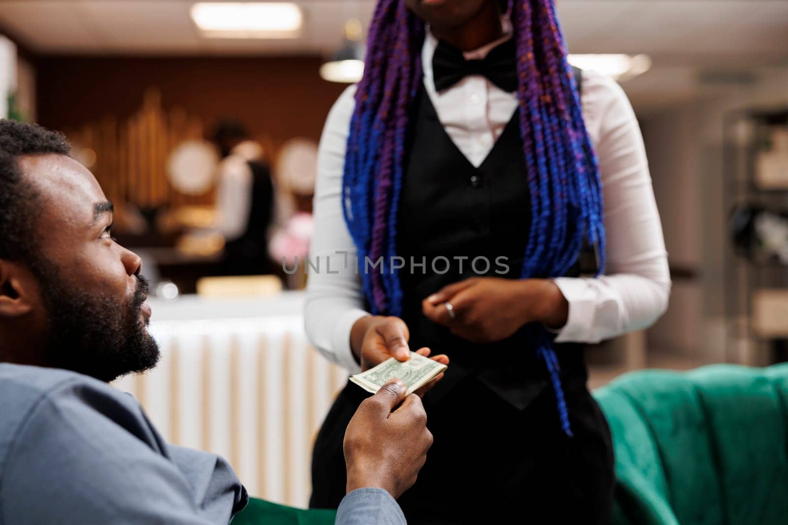 Young African American man hotel guest giving cash to waitress while relaxing in lobby, ordering food and drinks, tipping hospitality staff while resting at resort. Tipping behaviour in hospitality