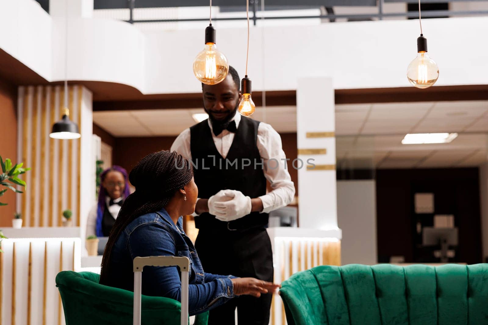 Smiling friendly hotel bellboy wearing uniform talking with African American woman guest sitting in lobby with luggage. Bellman greeting tourist, offering traveler to help carry suitcase
