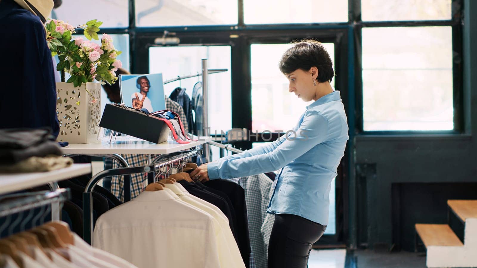 Store employee arranging hangers full with stylish clothes, working at modern boutique visual. Stylish worker putting fashionable merchandise on racks in shopping centre. Fashion concept