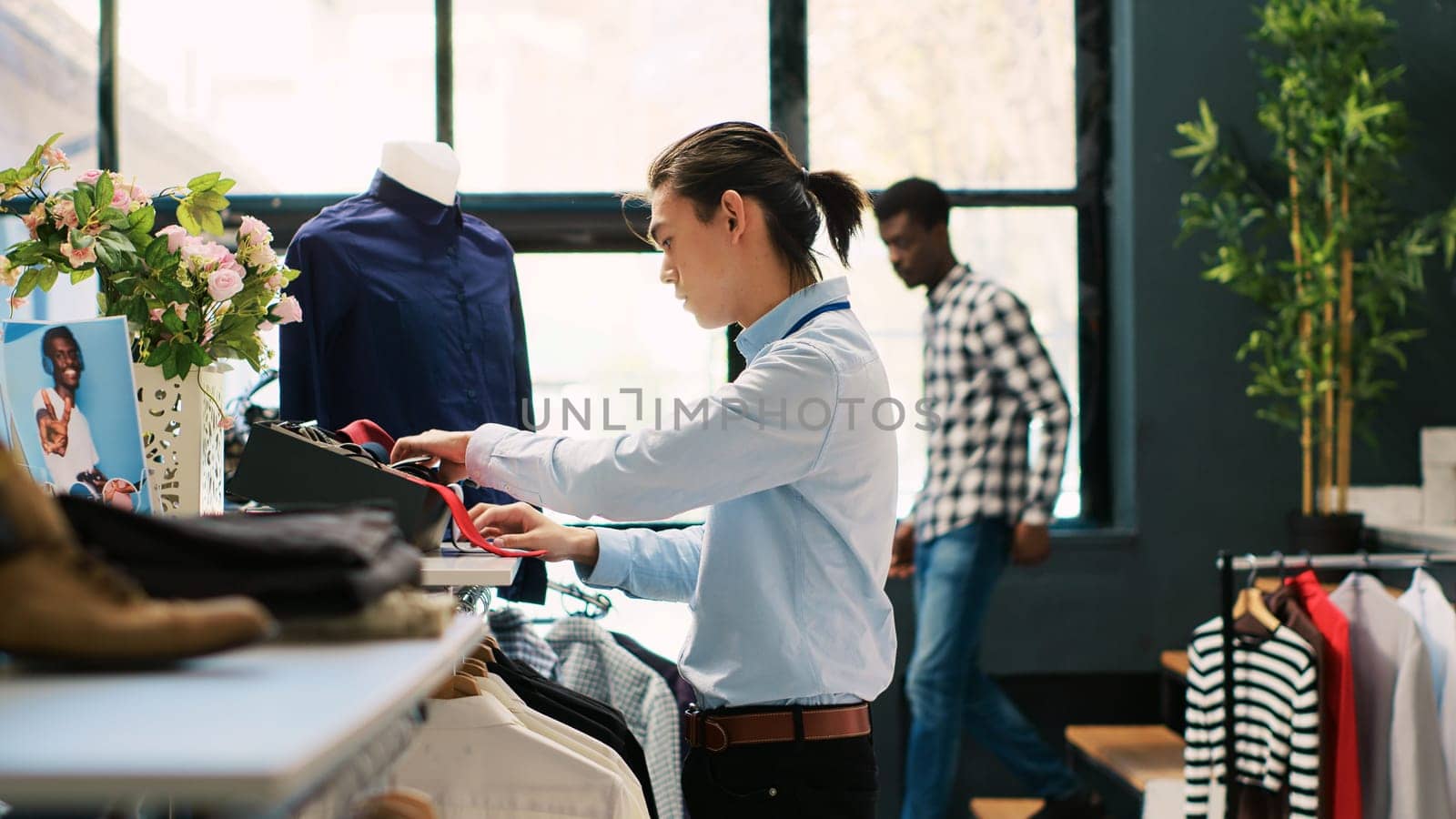 Asian worker arranging stylish tie, putting accessories on shelf in modern boutique. Retail employee preparing store for opening, working with fashionable clothes in shopping mall. Fashion concept