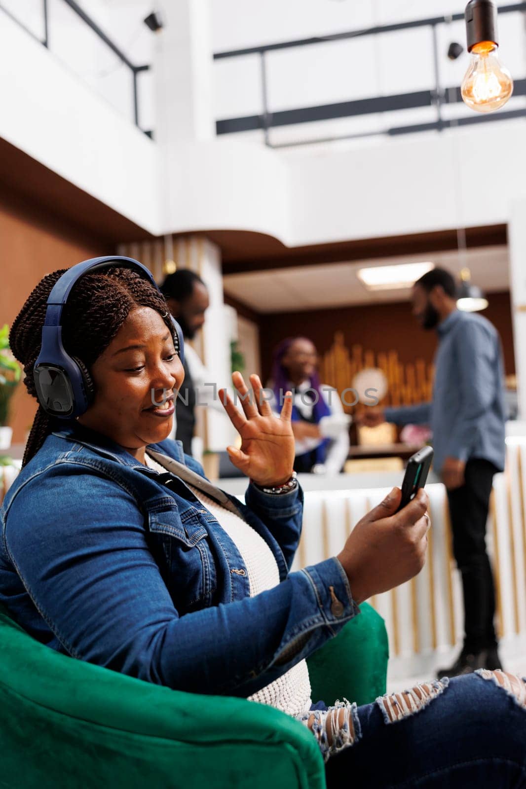 Happy African American young woman traveler wearing headphones looking at smartphone screen waving hand while sitting in hotel lobby. People staying in touch with friends and family while traveling