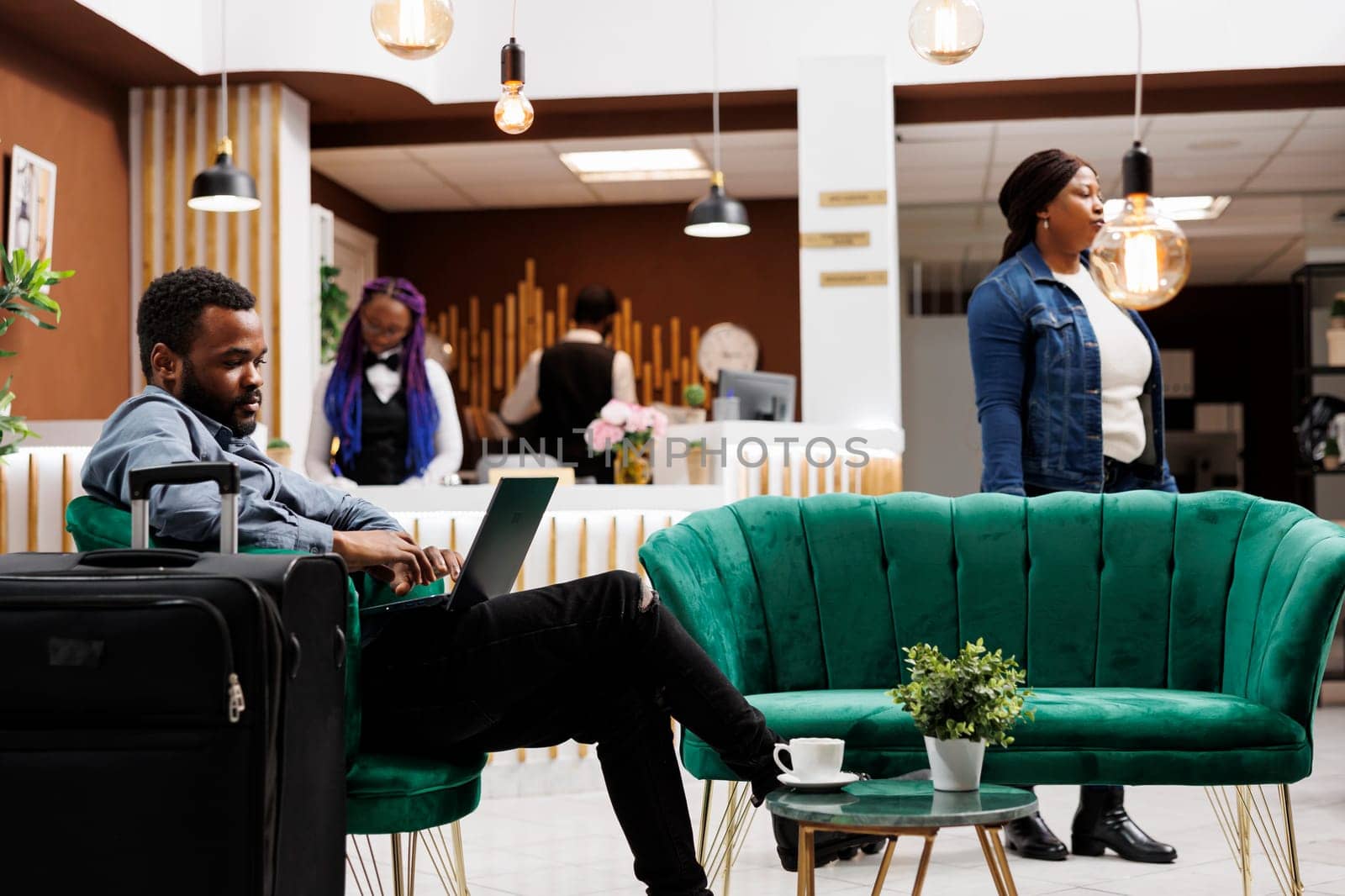 Busy African American man travelling freelancer sitting in armchair with suitcase and laptop at hotel lobby. Black millenial guy using computer working on project remotely during business trip