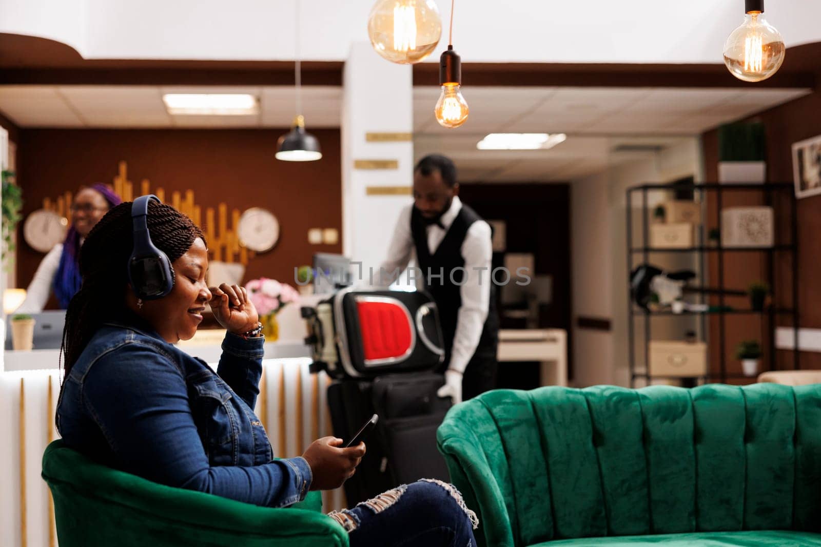 Happy African American woman tourist in headphones resting in hotel lobby listening to music on smartphone, waiting for check-in, arrived at tropical resort. Bellboy carrying baggage on background