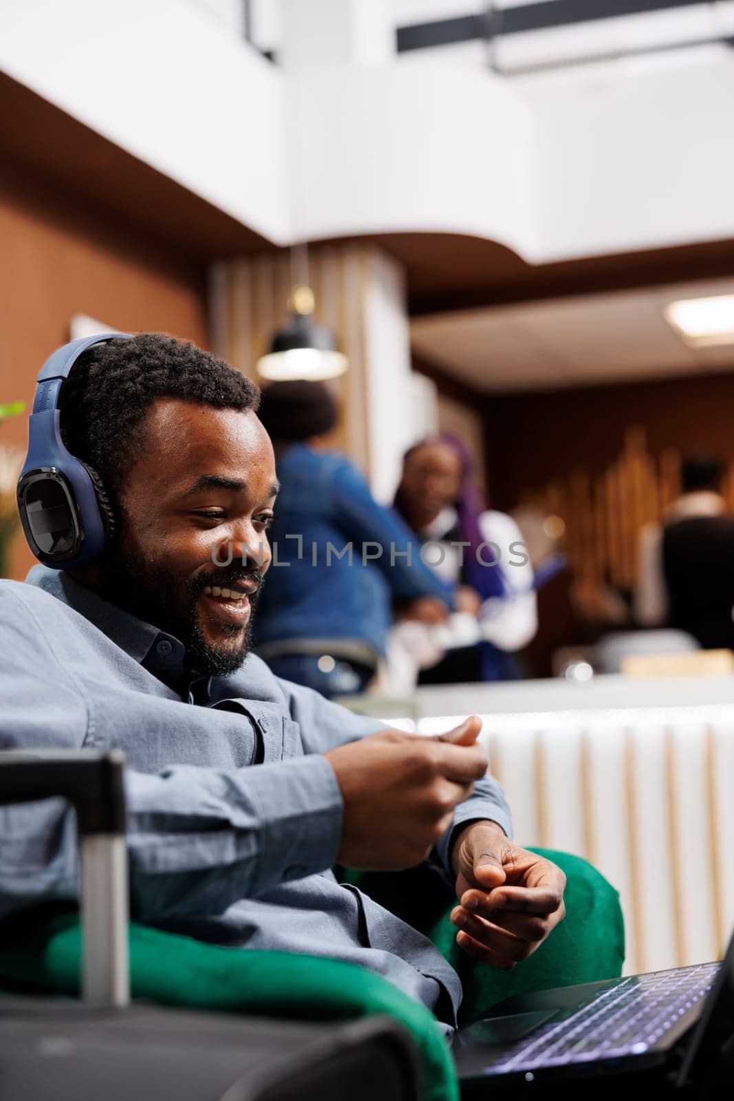 African American businessman wearing headphones using laptop for video call, waiting in hotel lounge area for check-in. Smiling black guy entrepreneur participating in virtual meeting while traveling