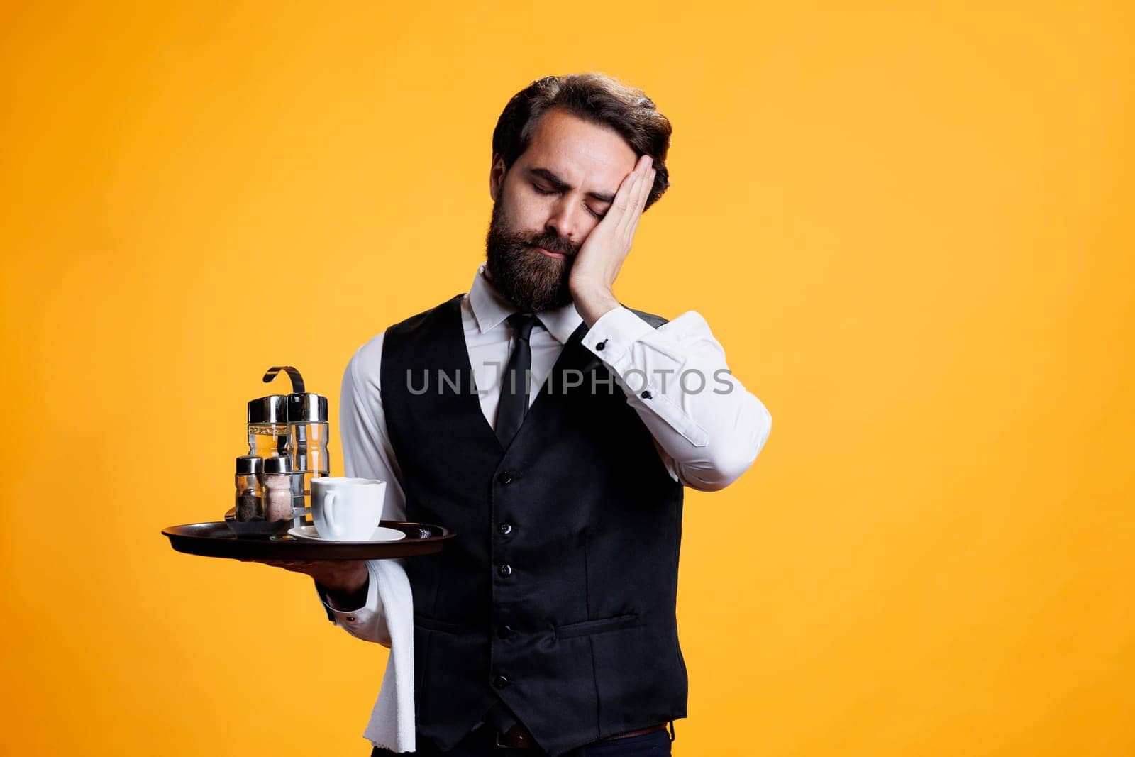 Young man dealing with burnout on camera, falling asleep while he stands against yellow background. Elegant waiter dressed with suit and tie feeling exhausted and overworked in studio.