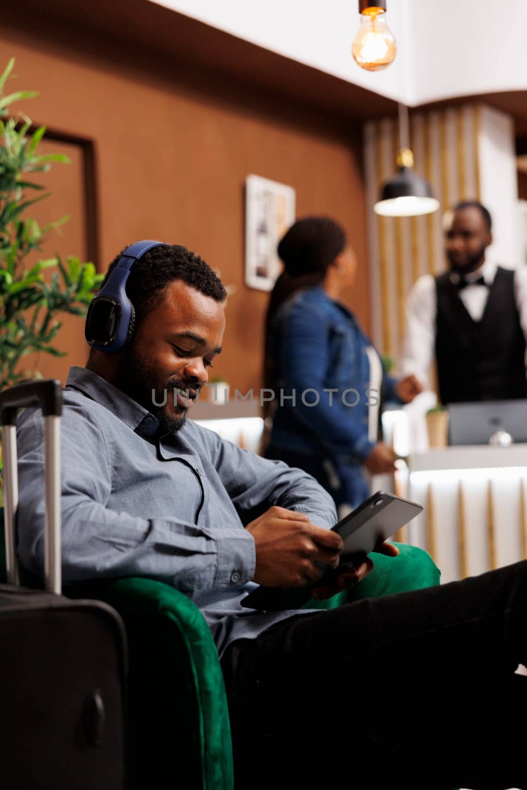 Smiling African American guy traveler sitting with luggage in hotel lobby, male tourist holding digital tablet connecting internet wifi while waiting for room to be ready. Guest self check-in