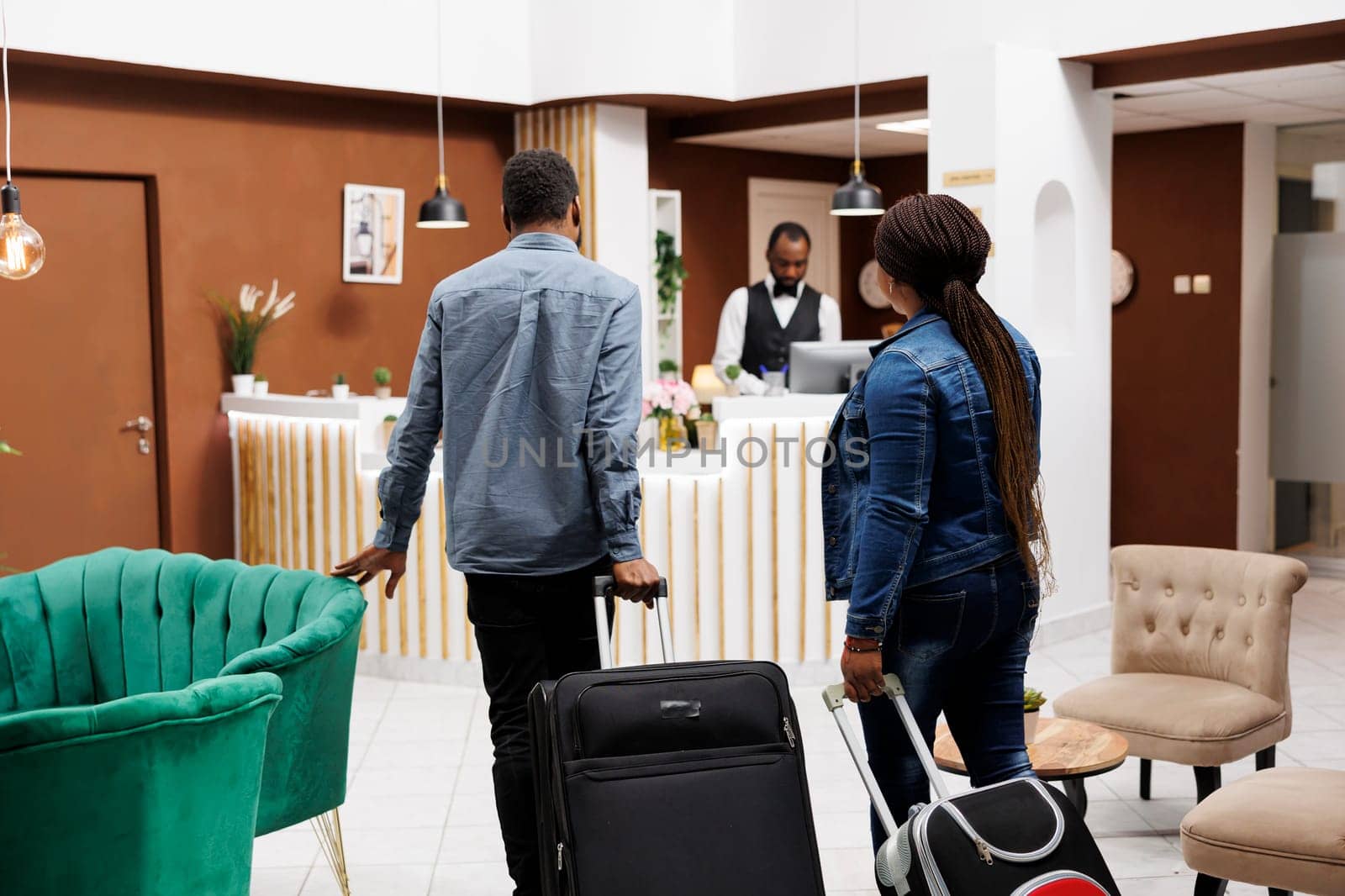 Rear view of young African American family couple arriving to resort hotel, people tourists pulling luggage going to reception desk to check-in. People, travel and vacation concept