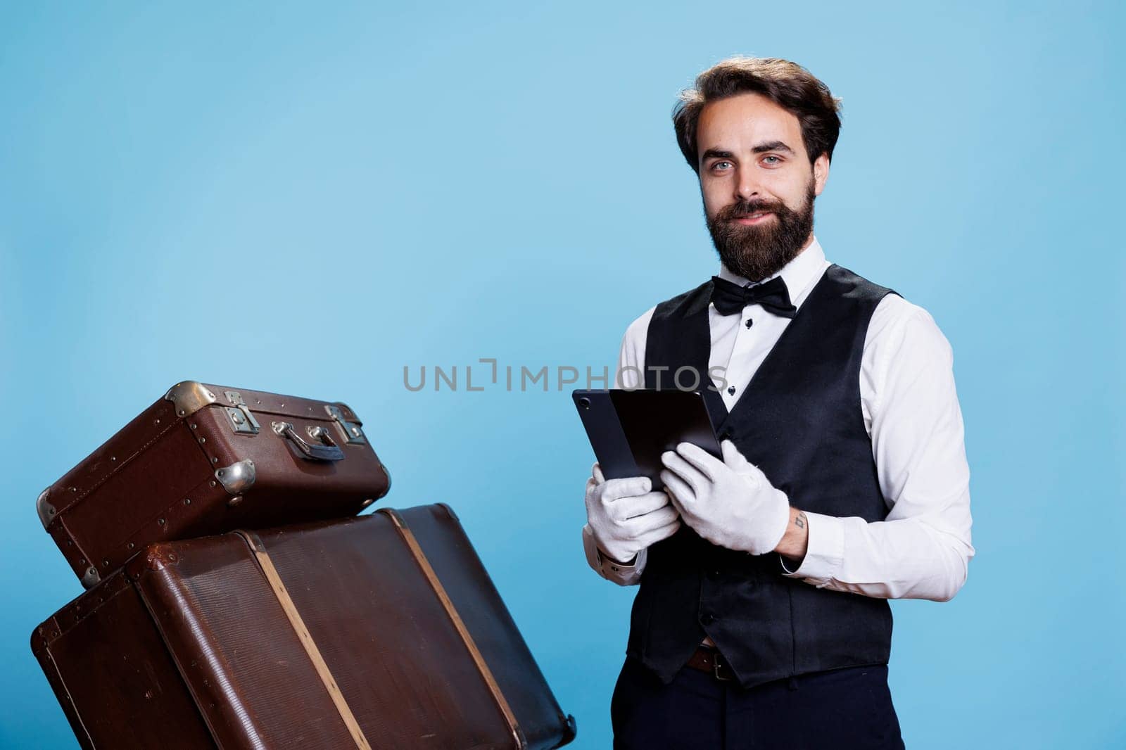 Hotel concierge checks online reservations on gadget, standing next to suitcases for safekeeping. Bellhop showcases his commitment in providing excellent services, important role.